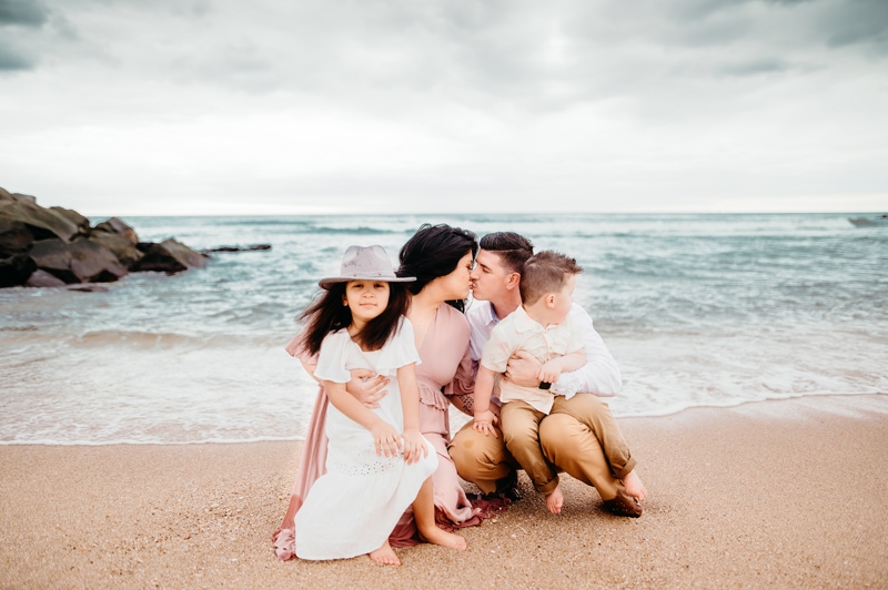 Family Photographer, mom and dad kiss as they hold their two children in their arms at the beach